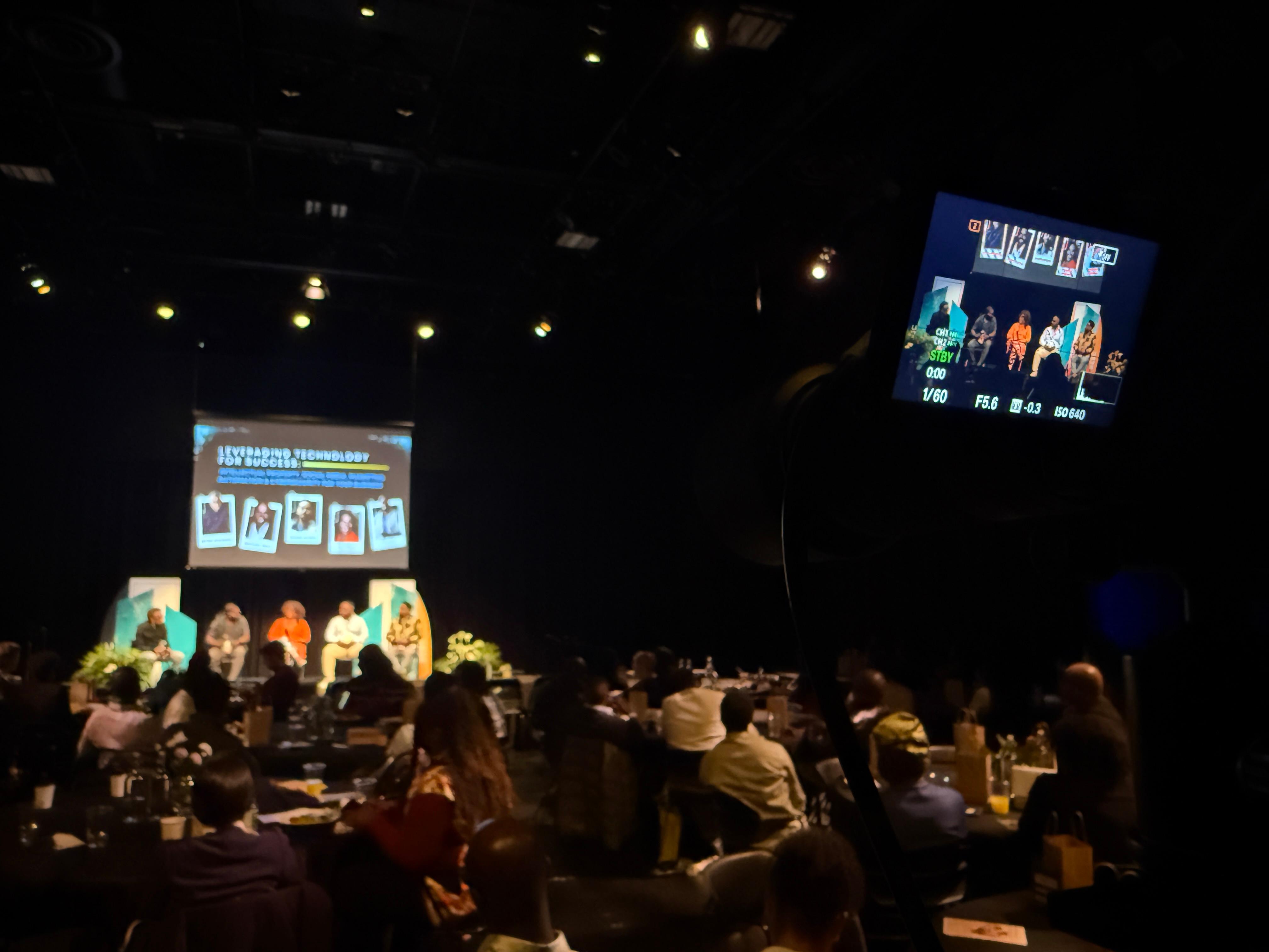 Event videography setup capturing a live speaker on stage with slides projected behind and engaged audience watching attentively.